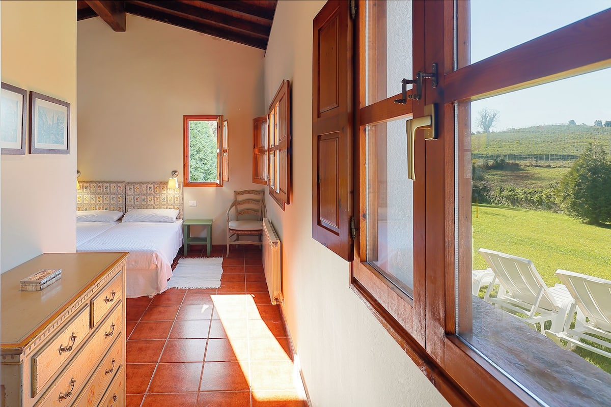 A bedroom with a bed, dresser, chair, and a view of a green landscape through a window.