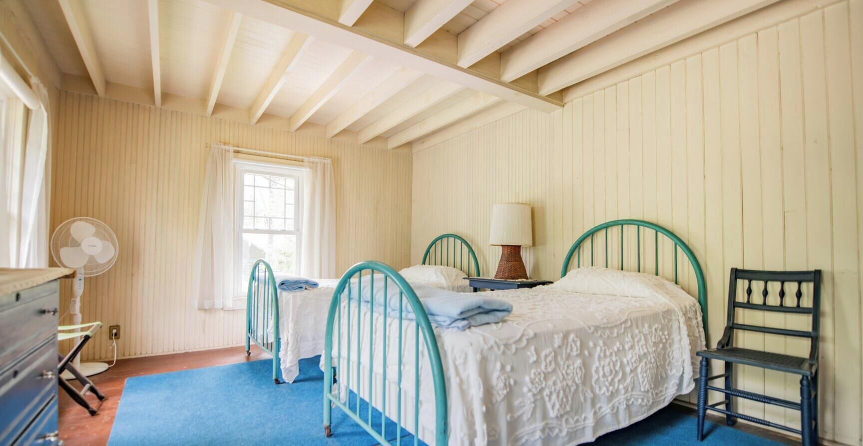 A bedroom with two single beds, a chair, a fan, and a window.