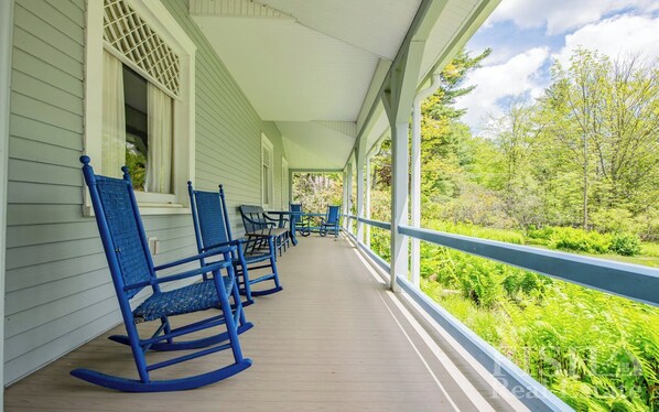 A covered porch with blue rocking chairs and a railing overlooking a lush green landscape.