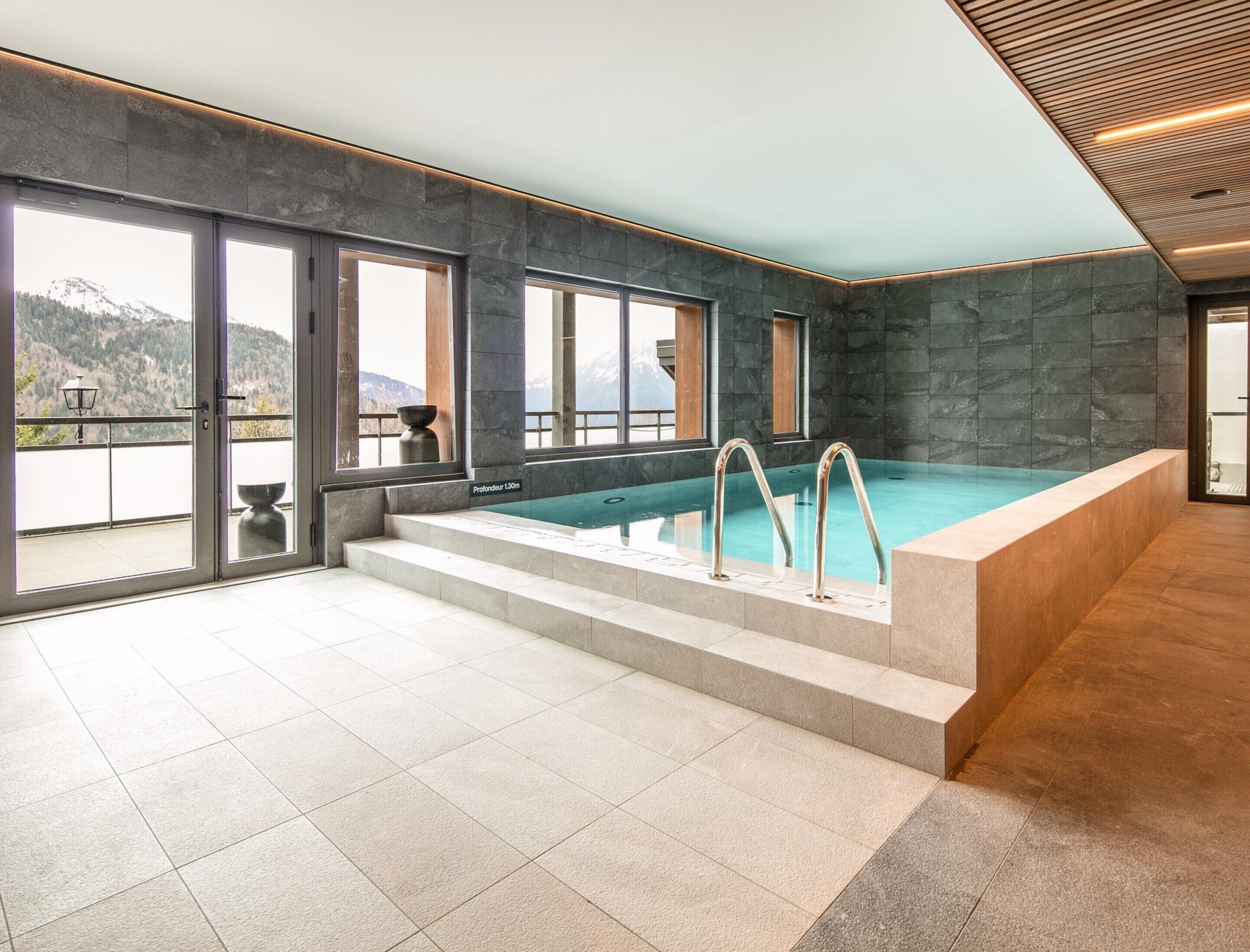 En modern inomhuspool med utsikt över berg, stenmurar och en balkong.