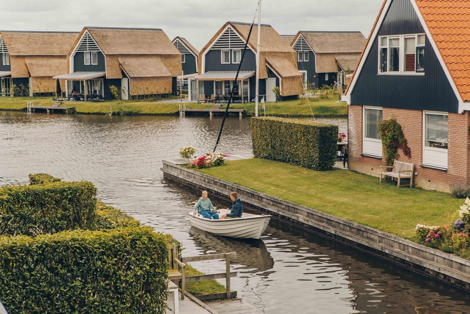 Huse rækket op langs en kanal, en båd med to personer, og et grønt område med en bænk.