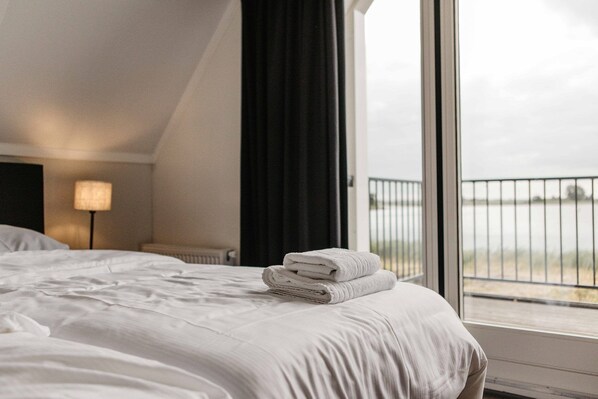 A hotel room with a bed, a lamp, a window with a view of a body of water, and a balcony.