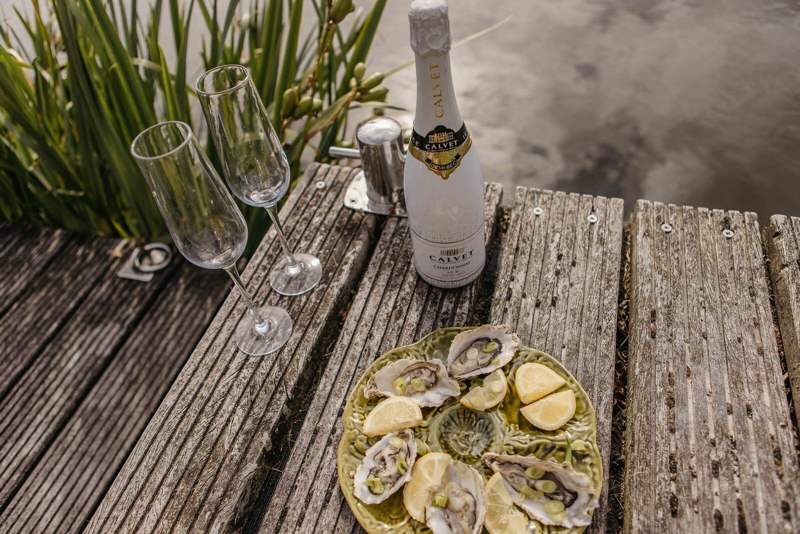 Une table en bois sur laquelle se trouvent une bouteille de Calvet Chardonnay, deux verres vides et une assiette d’huîtres garnie de tranches de citron.