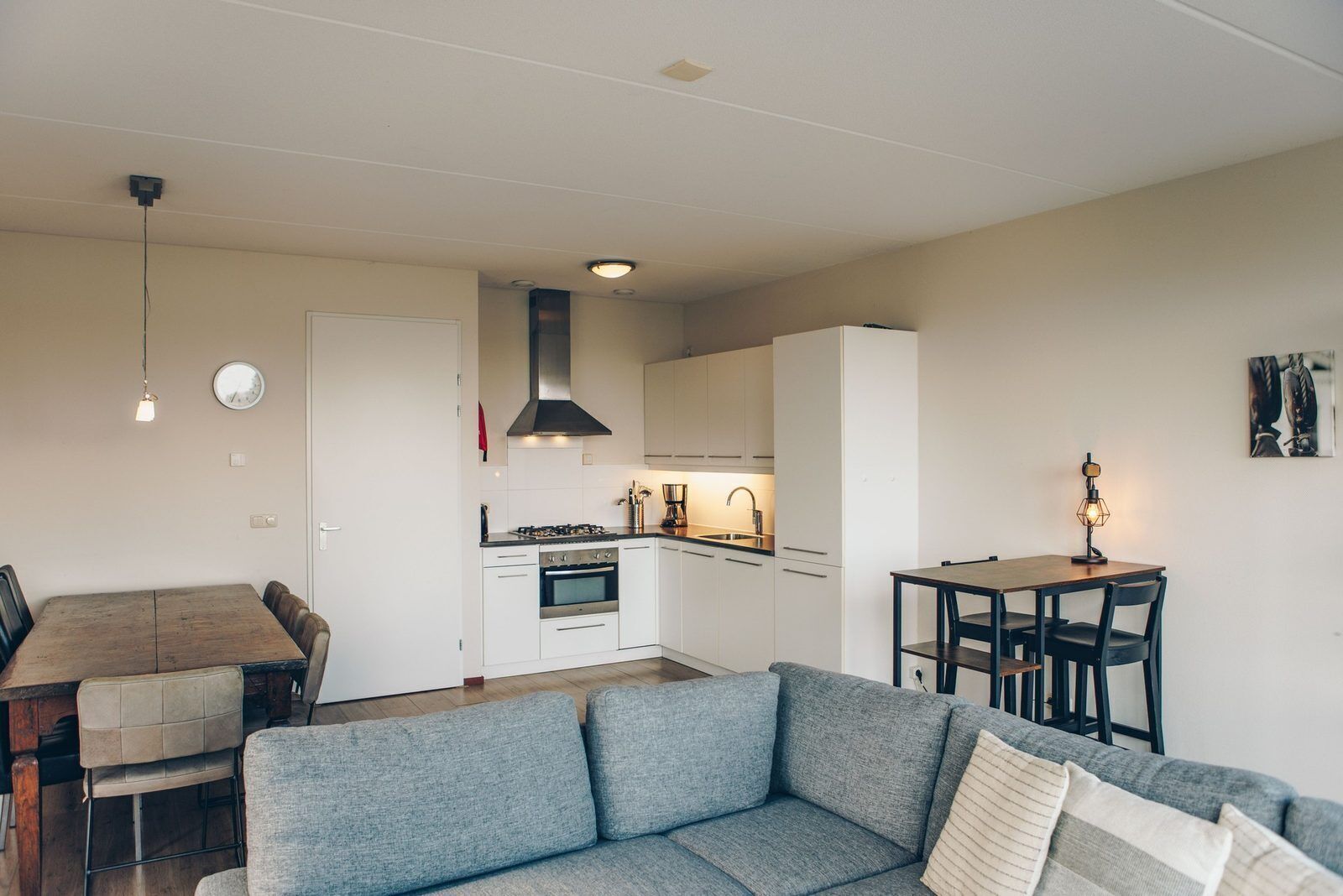 Une cuisine moderne avec des meubles blancs, une cuisinière et un évier. On y trouve un coin repas avec une table et des chaises en bois, ainsi qu’un coin salon avec un canapé gris.
