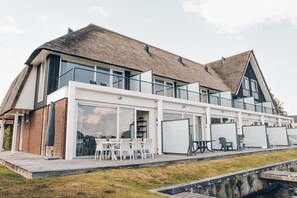 Une maison moderne dotée d’une grande façade vitrée, d’un toit de chaume et d’un balcon aménagé avec des sièges d’extérieur.