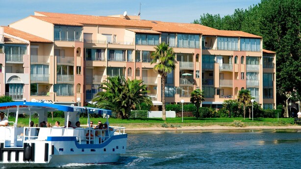 A multi-story hotel with balconies, a boat docked in front, and palm trees.
