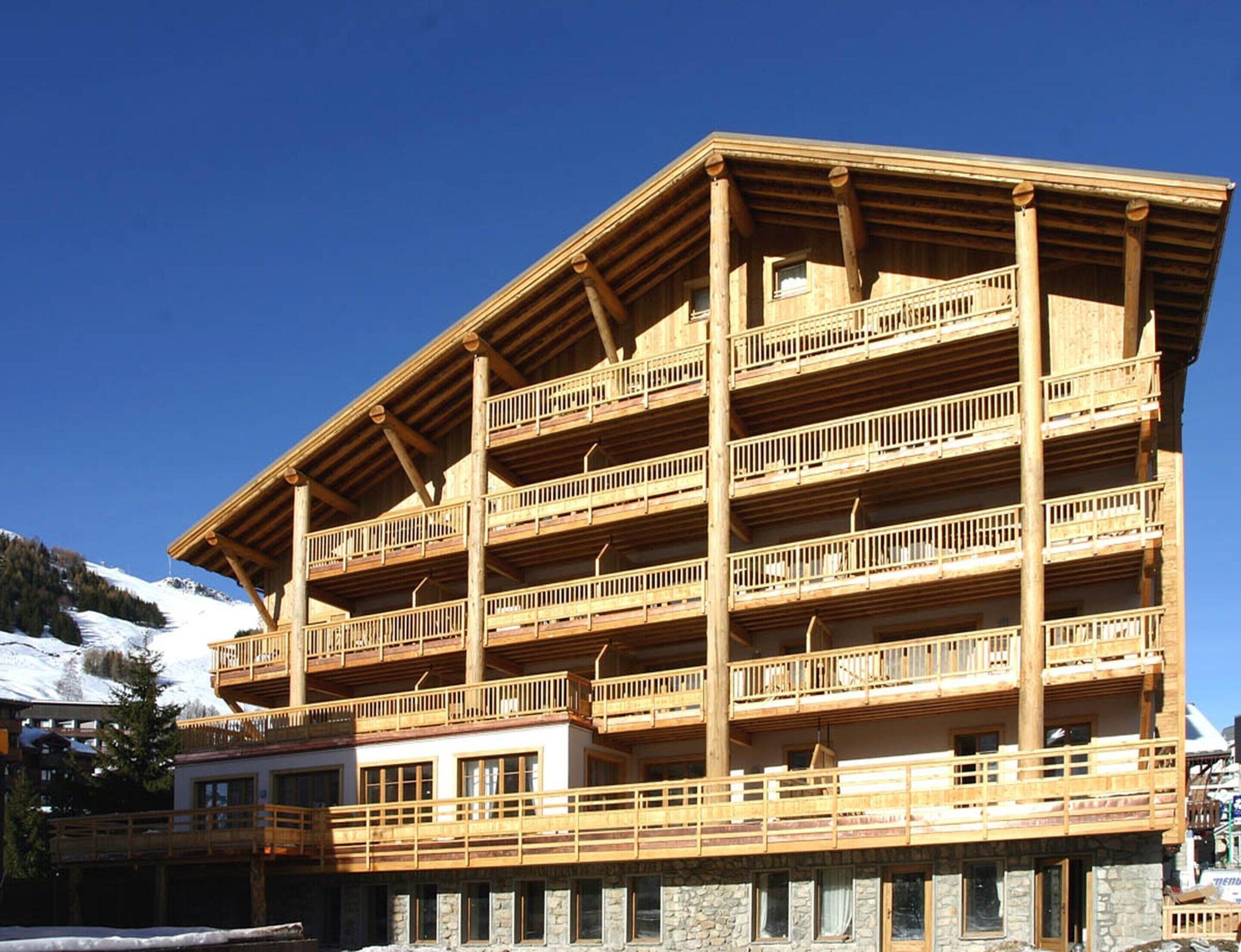 A multi-story wooden building with balconies, likely a hotel or lodge, with a clear sky and snow in the background.