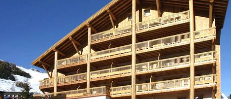A multi-story wooden building with balconies, likely a hotel or lodge, with a clear sky and snow in the background.