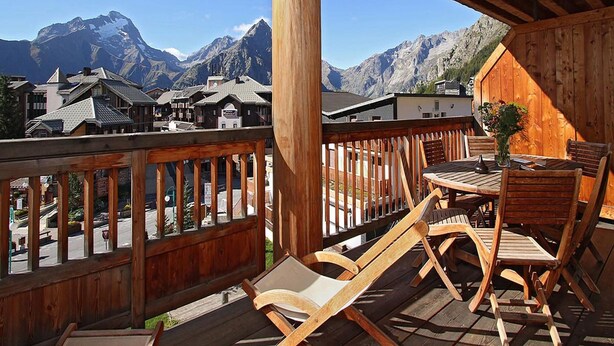 Une terrasse en bois avec une table et des chaises, offrant une vue sur un paysage montagneux parsemé de bâtiments.