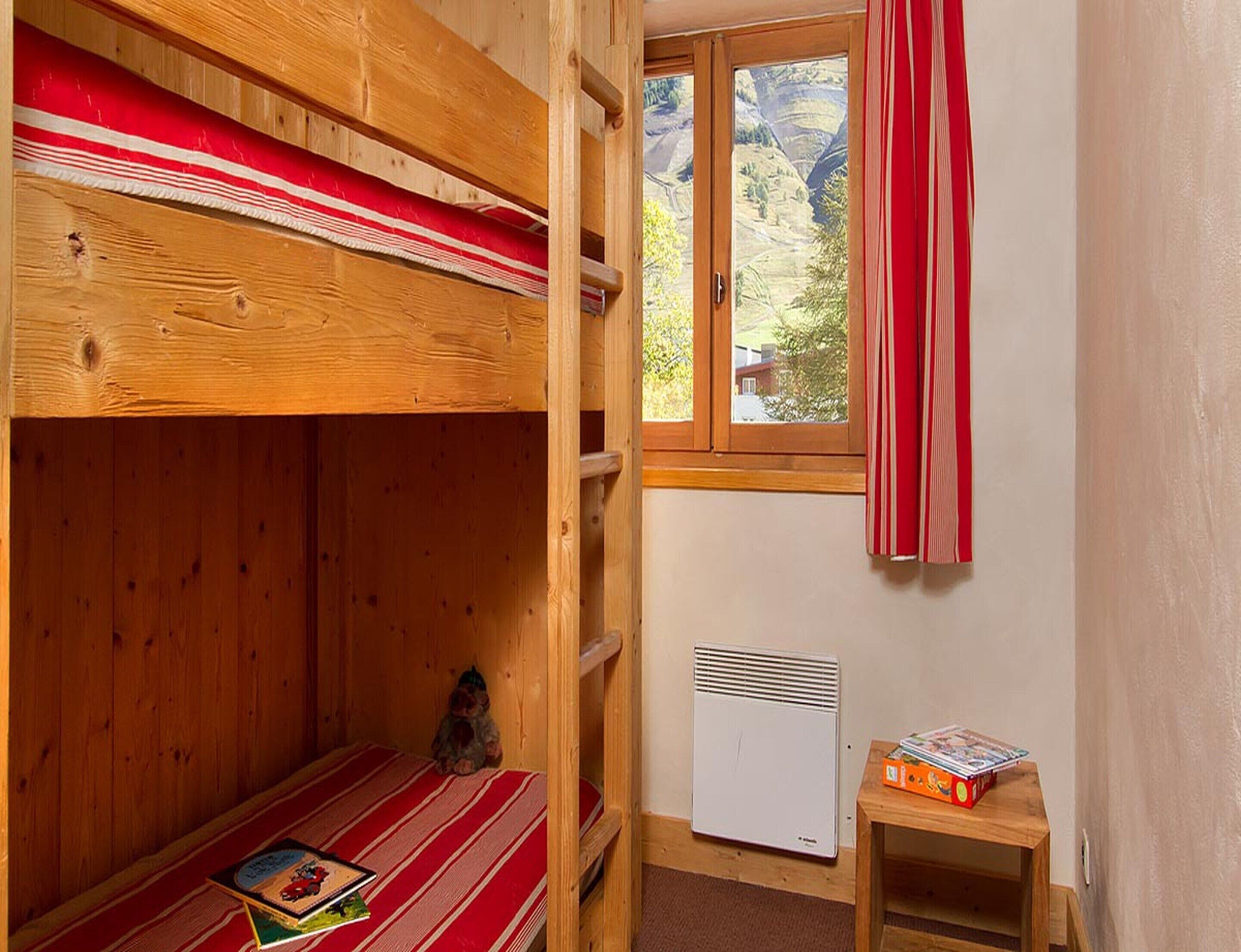 A bunk bed with a red and white striped bedspread, a small table with books, a radiator, and a window with a view of a mountainous landscape.