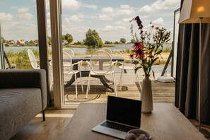 A room with a laptop on a table, a sofa, and a view of a lake with chairs outside. - Parc Pharshoeke - Herenhuis geschakeld | 6p (Heeg)