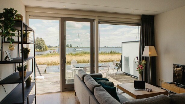 A modern living room with a sofa, a coffee table, a TV, and a bookshelf. There is a sliding glass door leading to an outdoor area with a view of the water and a dock. - Parc Pharshoeke - Herenhuis geschakeld | 6p (Heeg)