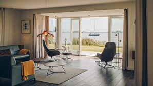 A modern living room with a sofa, chairs, a table, and a view of a lake with a boat. - Parc Pharshoeke - Meervilla Deluxe met Sauna | 8p (Heeg)