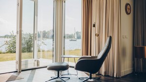 A room with a view of a lake, a chair, a table, and a curtain. - Parc Pharshoeke - Meervilla Deluxe met Sauna | 8p (Heeg)