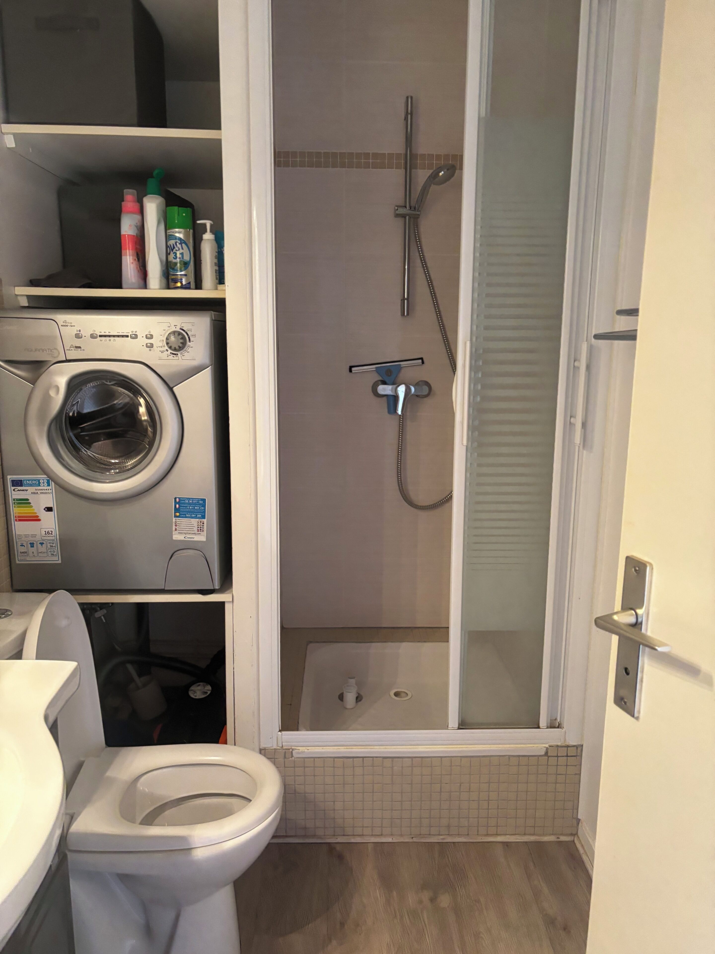 Une petite salle de bain équipée d’un lave-linge, de toilettes et d’une douche séparée par une paroi en verre.