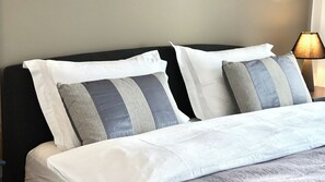 Una cama bien hecha con almohadas a rayas blancas y grises, un cabecero oscuro y una lámpara en una mesita de noche.