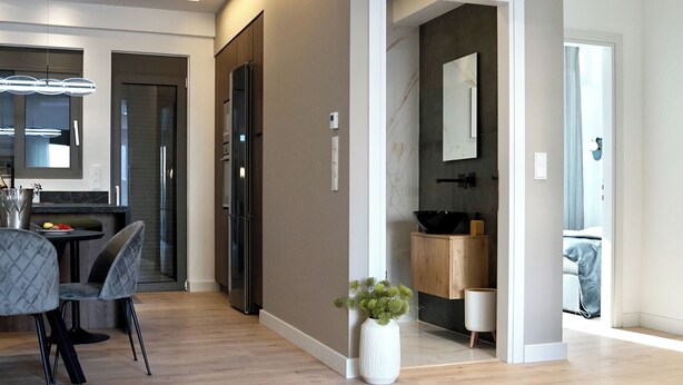 A modern kitchen with dark cabinetry, a dining area with a table and chairs, and a hallway leading to a bathroom with a large mirror and a sink. - Alimos Escape 2 (Alimos)