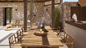 Une table à manger en bois avec des chaises, un mur en pierre et un panier suspendu rempli de fruits.