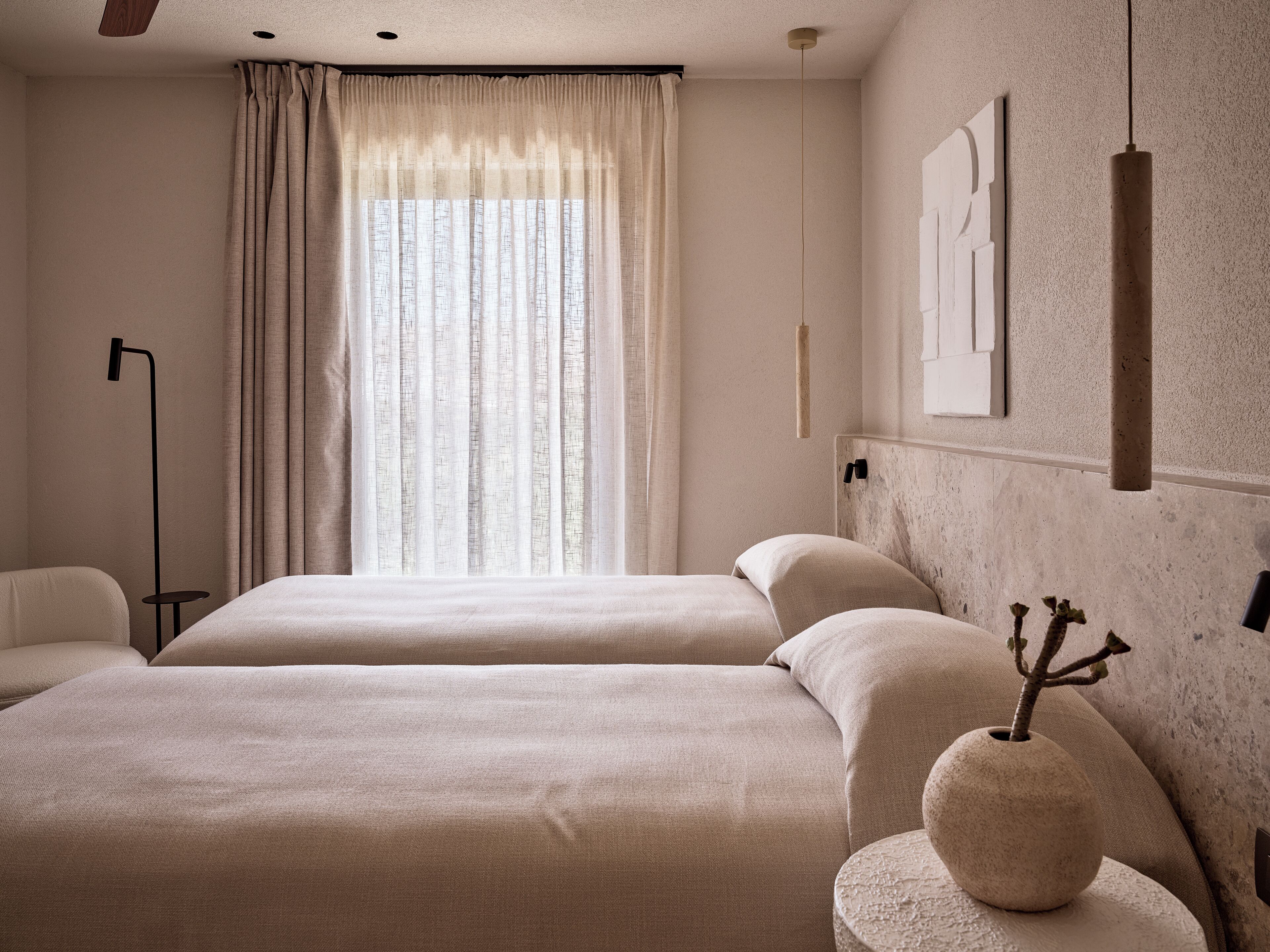 A hotel room with two beds, a large window with curtains, a floor lamp, and a vase with branches on a side table.