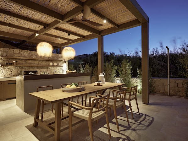 An outdoor dining area with a stone wall, wooden table, and chairs. - Villa Alemar - Delmar Collection (Milos)