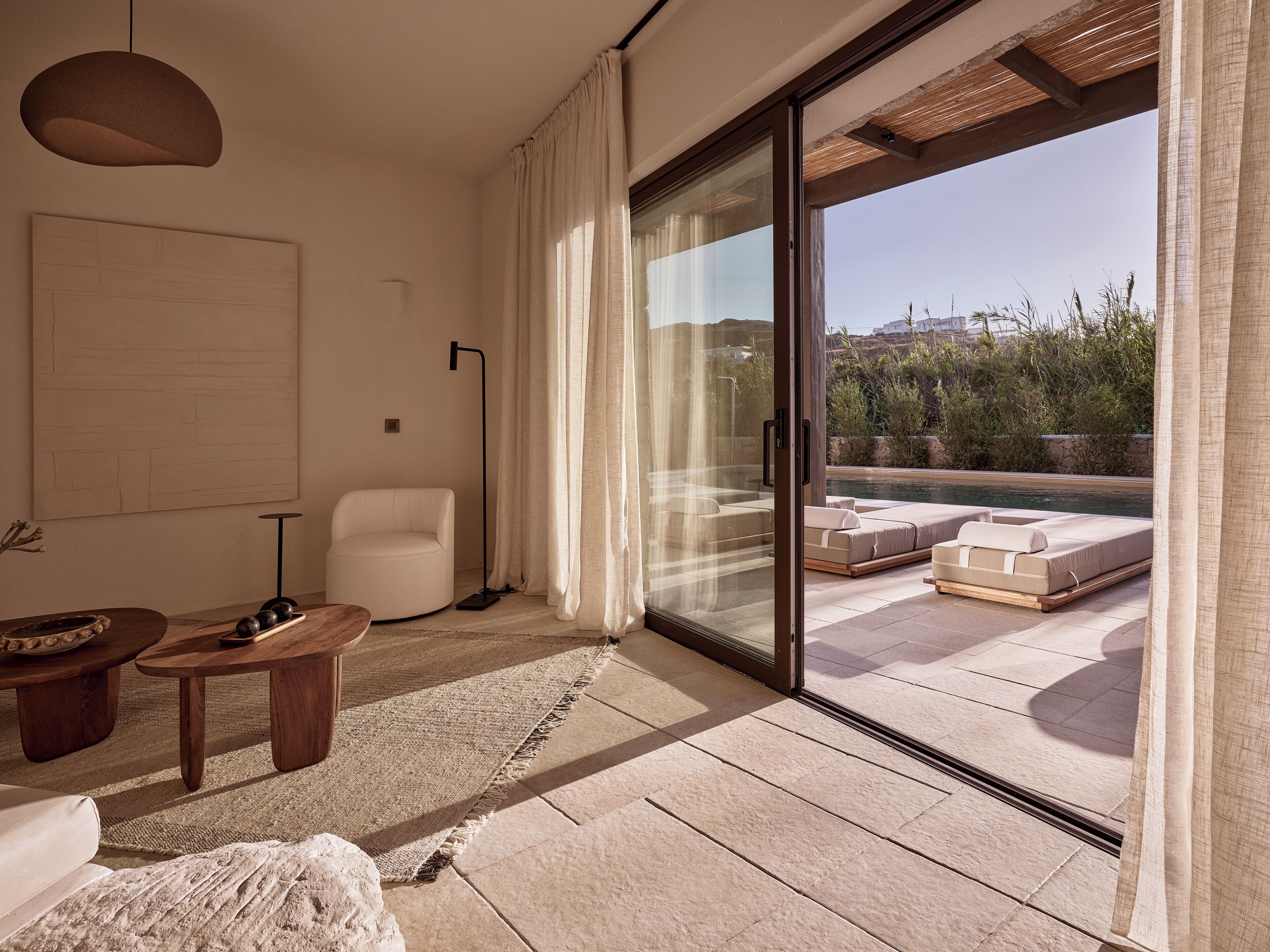 A modern living room with a sofa, a coffee table, and a floor lamp. There is a sliding glass door leading to a balcony with a pool and outdoor furniture.