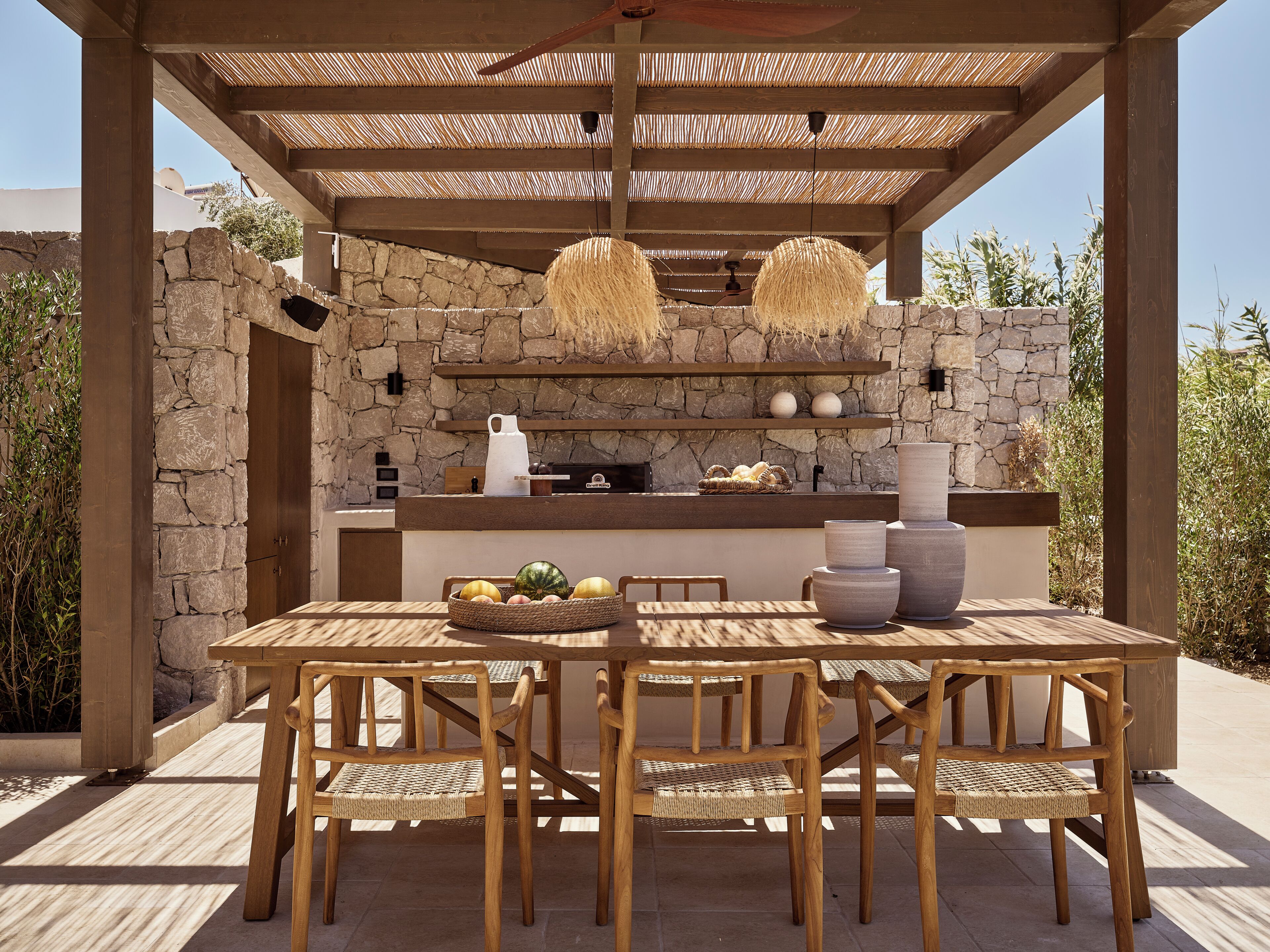 An outdoor dining area with a stone wall, wooden pergola, and woven chairs.