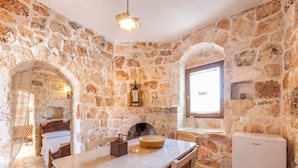 Una habitación con paredes de piedra, una cama, una mesa con sillas, un lavamanos y una ventana.