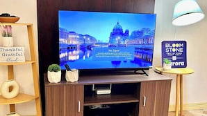 Un televisor mostrando un paisaje urbano, un mueble de madera para equipo electrónico, una mesita con una lámpara y un cartel con el texto 'BLOOM STONE'.