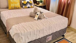 Una cama bien hecha con una manta color beige, dos almohadas, una toalla enrollada y una mesita de noche con una lámpara y una pequeña planta.