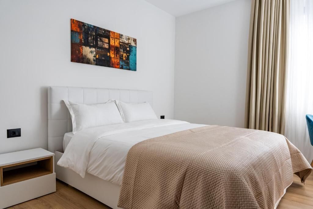 Una habitación de hotel moderna con una cama blanca, una mesita de noche pequeña y un cuadro abstracto colorido en la pared.