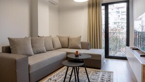 A modern living room with a sofa, a small round table, a hanging light fixture, a sliding glass door to a balcony, and a view of a cityscape. - Barrikada - E Apartments (Tirana)