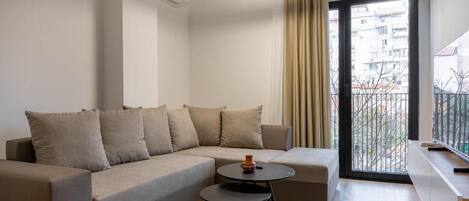 A modern living room with a sofa, a small round table, a hanging light fixture, a sliding glass door to a balcony, and a view of a cityscape.