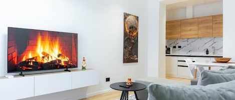 Una sala de estar moderna con chimenea, televisor de pantalla plana, sofás grises, una mesita redonda y una cocina al fondo.