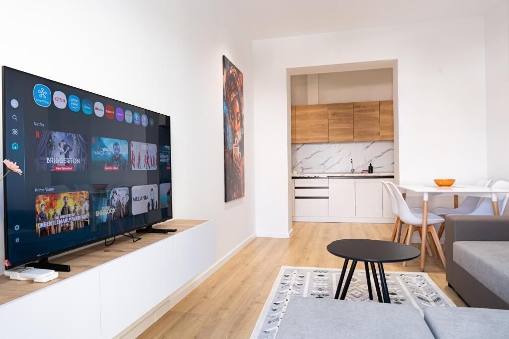 A modern living room with a flat-screen TV displaying streaming services, a dining area with a white table and chairs, and a kitchen with wooden cabinets and a marble backsplash.