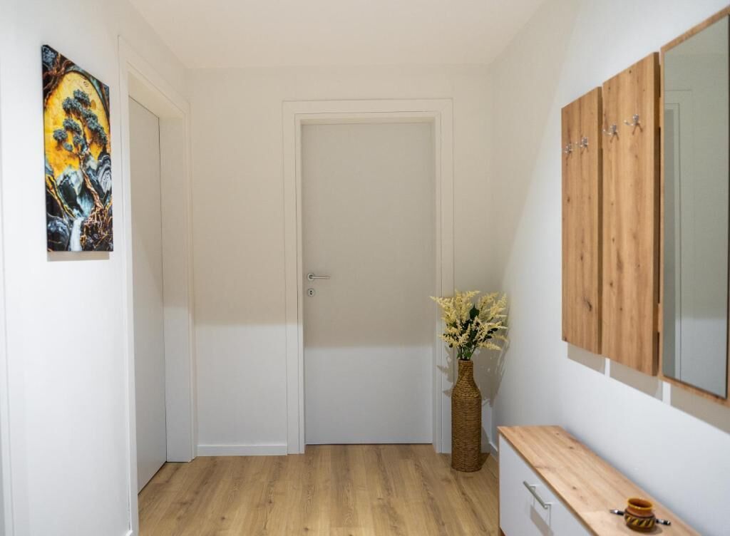 Un couloir avec une porte blanche, un banc en bois, un vase de fleurs et un tableau accroché au mur.