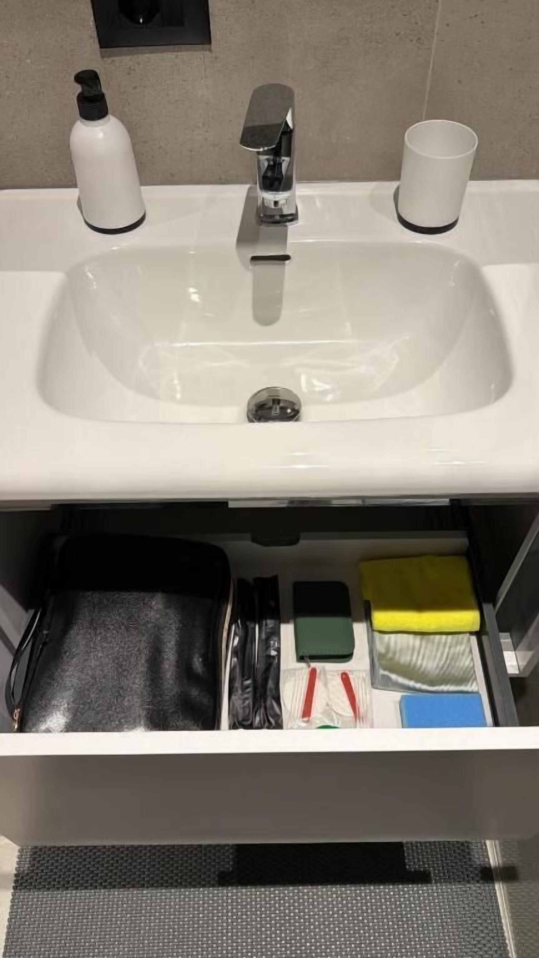 Ett handfat på hotellrummets badrum, med en spegel ovanför, en handduksställning och en förvaringslåda under som innehåller toalettartiklar och en svart väska.