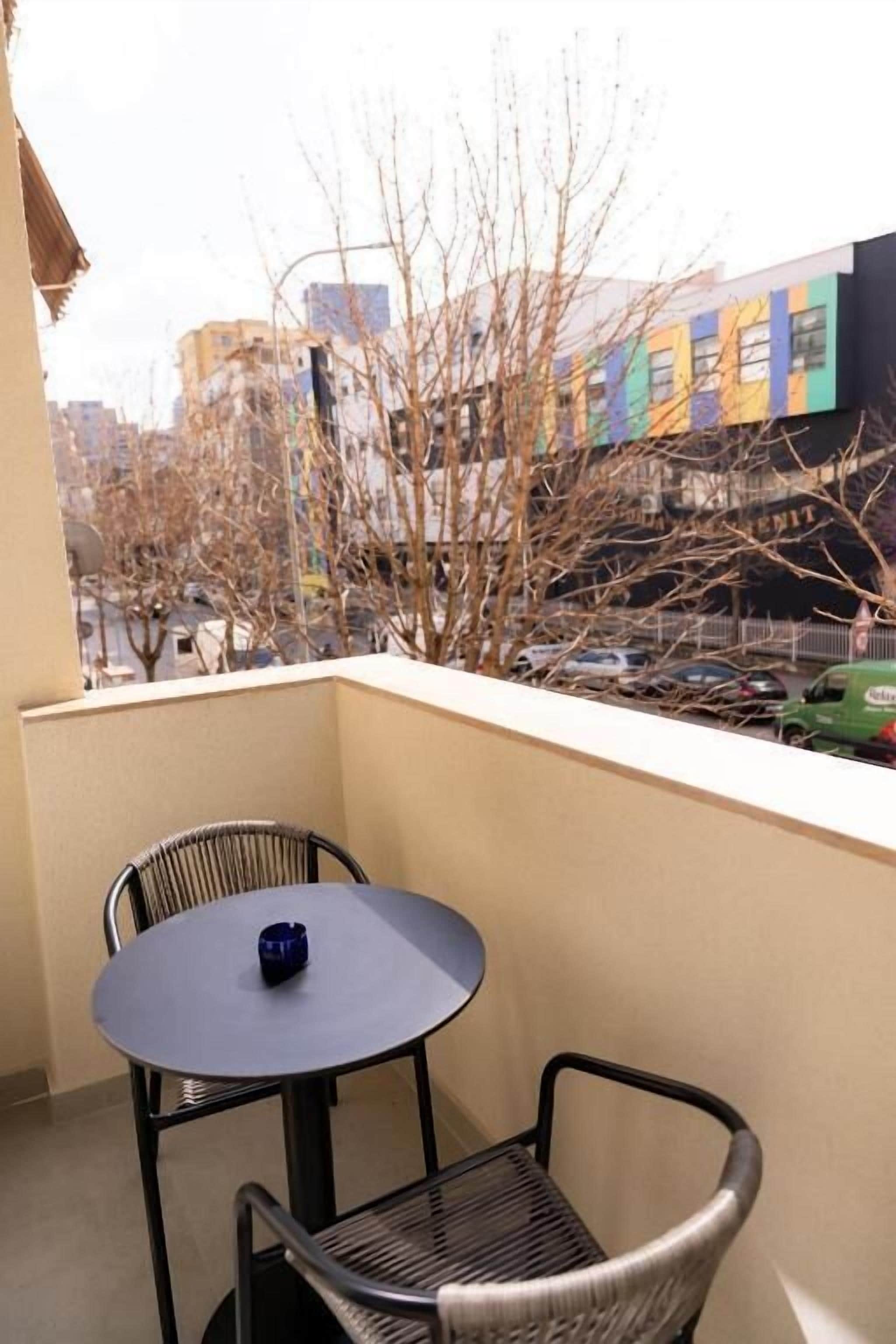 A balcony with a round table and two chairs, overlooking a street with buildings and parked cars.