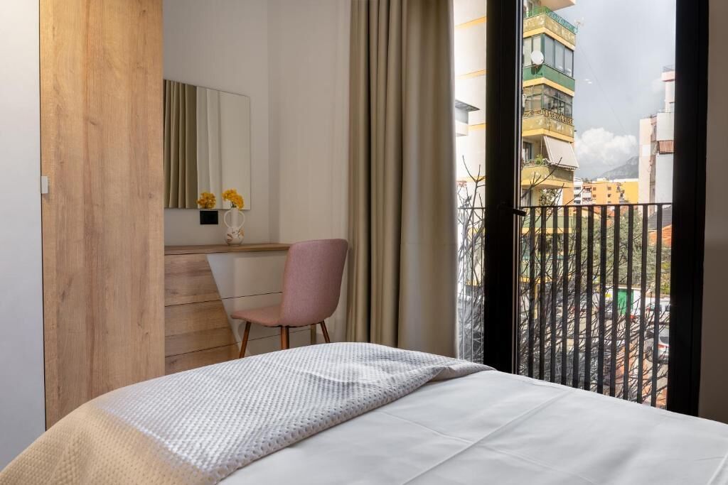 Ein Schlafzimmer mit einem Bett, einem Schreibtisch, einem Stuhl, einem Spiegel und Blick auf Gebäude durch ein Fenster.