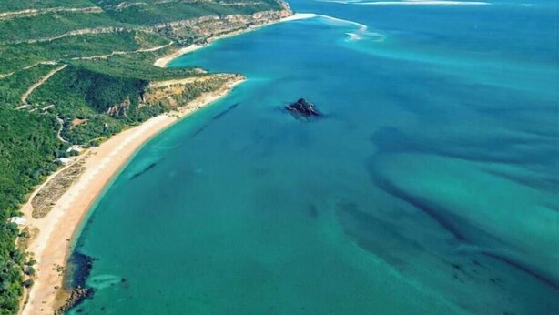 Vista aérea de una zona costera con playa, aguas azules cristalinas y colinas verdes.