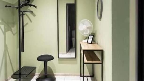 Ein Zimmer mit Fliesenboden, einem Kleiderhaken, einem kleinen Hocker, einem Spiegel und einem Ventilator auf einem Tisch.