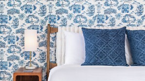 Ein Schlafzimmer mit gemustertem Wandpapier, einem Holfnachtkästchen, einem Nachttisch und einem Bett mit weißer Bettwäsche und blauen Kissen.