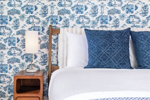 Ein Schlafzimmer mit gemustertem Wandpapier, einem Holfnachtkästchen, einem Nachttisch und einem Bett mit weißer Bettwäsche und blauen Kissen.