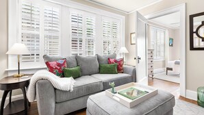 Ein Wohnzimmer mit grauem Sofa, grünen Kissen, einer weißen Tagesdecke, einem Beistelltisch mit Lampe und einem Tablett mit Büchern.