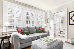 Ein Wohnzimmer mit grauem Sofa, grünen Kissen, einer weißen Tagesdecke, einem Beistelltisch mit Lampe und einem Tablett mit Büchern.