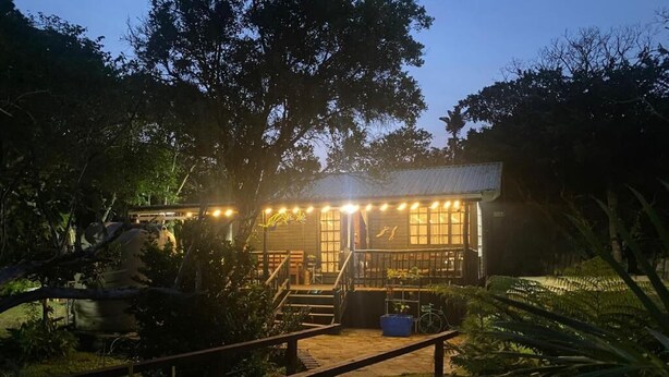A wooden house with a porch, surrounded by greenery, and a wooden bridge leading to it. - Little Creek Cabins (Port Edward)
