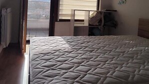 Un dormitorio con cama, ventana con persianas, radiador y vista a la ciudad.