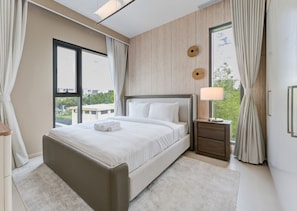 Une chambre moderne avec un grand lit, une table de chevet, une fenêtre avec des rideaux et une vue sur la verdure à l’extérieur.