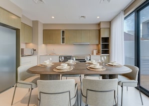 Un espace repas moderne avec une table ronde dressée pour six personnes, entourée de six chaises, dans une cuisine contemporaine dotée de meubles en bois et d’une grande fenêtre.