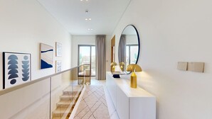 Passadís modern d'hotel amb una escala, una taula consola blanca amb una làmpada, un mirall gran i obra d'art a les parets.