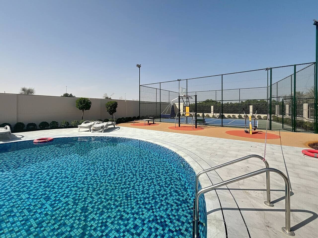 Une piscine avec un bord carrelé, un escalier en métal et une zone délimitée par une clôture, comprenant des bancs et un terrain de basketball.